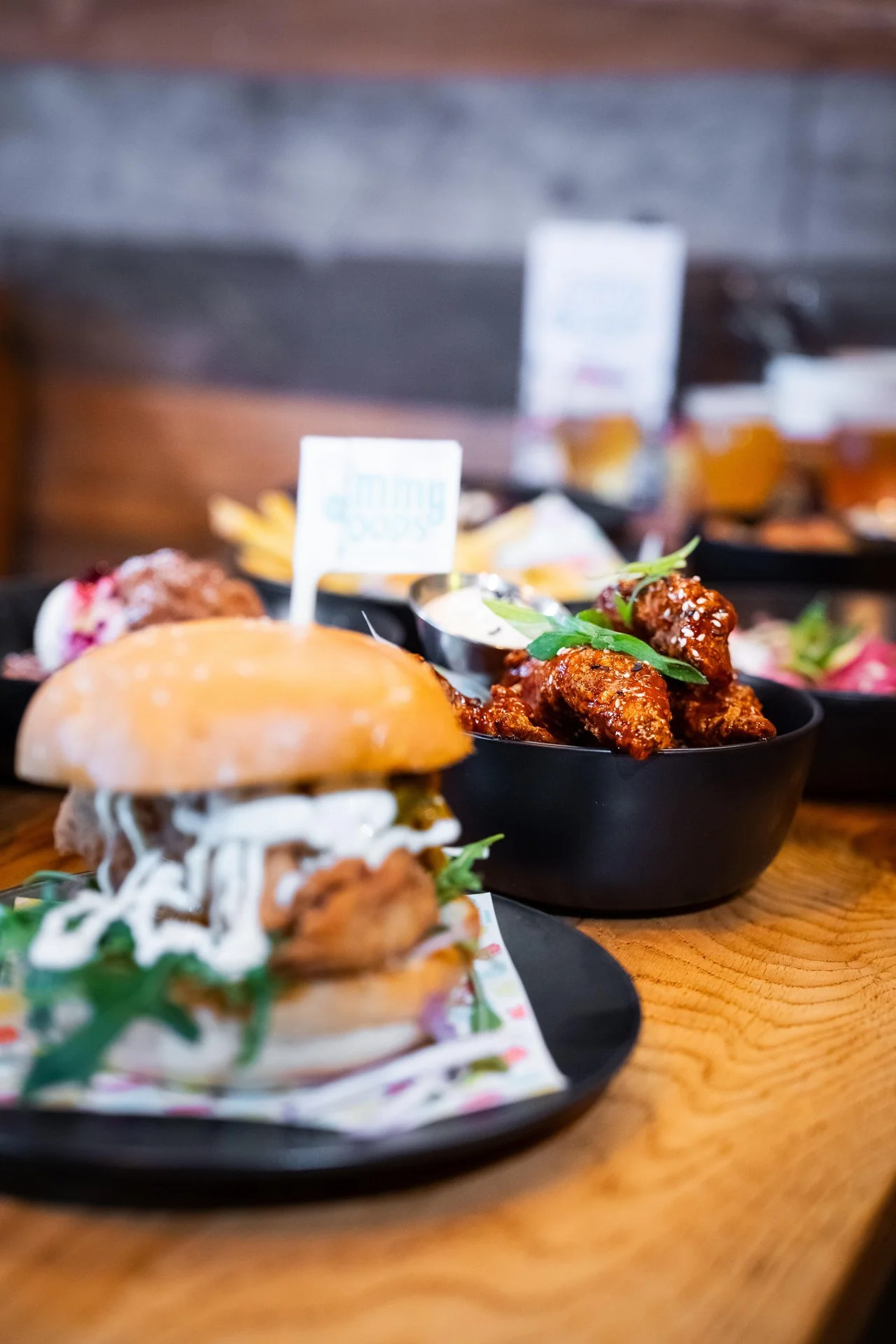 Close-up of a burger with fried chicken, lettuce, and sauce on a black plate, and a bowl of spicy glazed chicken wings garnished with green herbs on a wooden table.