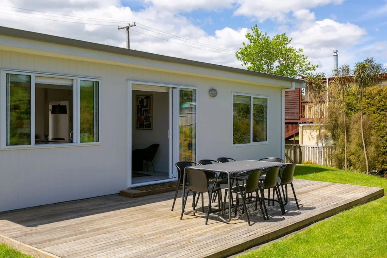 A backyard patio with a wooden deck, outdoor dining table, and six chairs in front of a white house with sliding glass doors