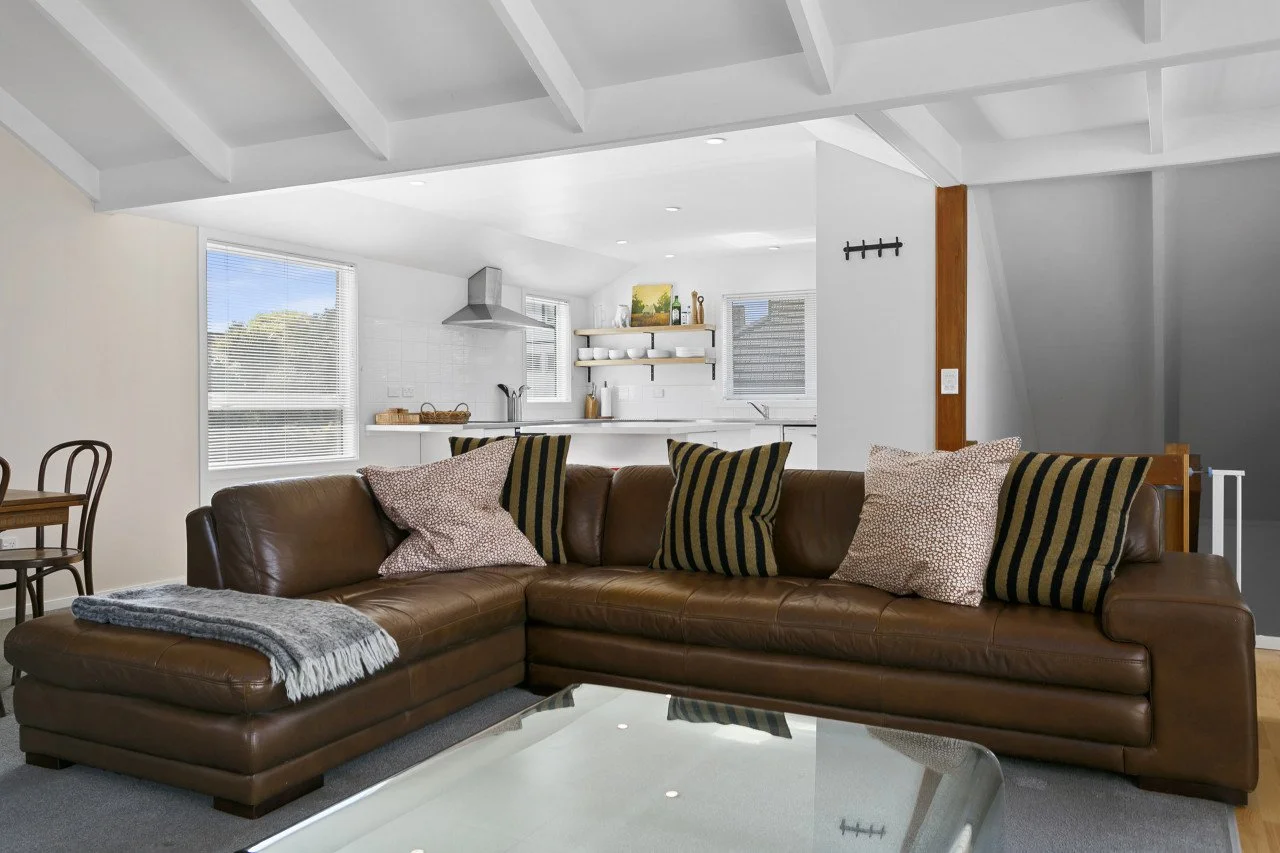 Living room with a brown leather sectional sofa, patterned pillows, a gray throw blanket, and a glass coffee table. In the background, an open kitchen with white cabinets, open shelves, a stainless steel range hood, and windows letting in natural lig
