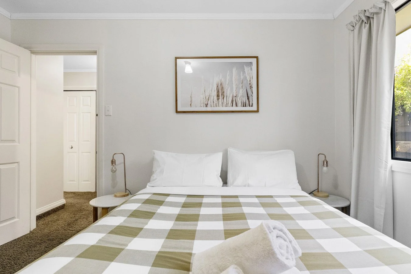 A neatly made bed with a beige and white checkered bedspread, two white pillows, rolled towels on the bed, flanked by two small nightstands with lamps, a framed artwork above the bed, and a window with white curtains to the right.