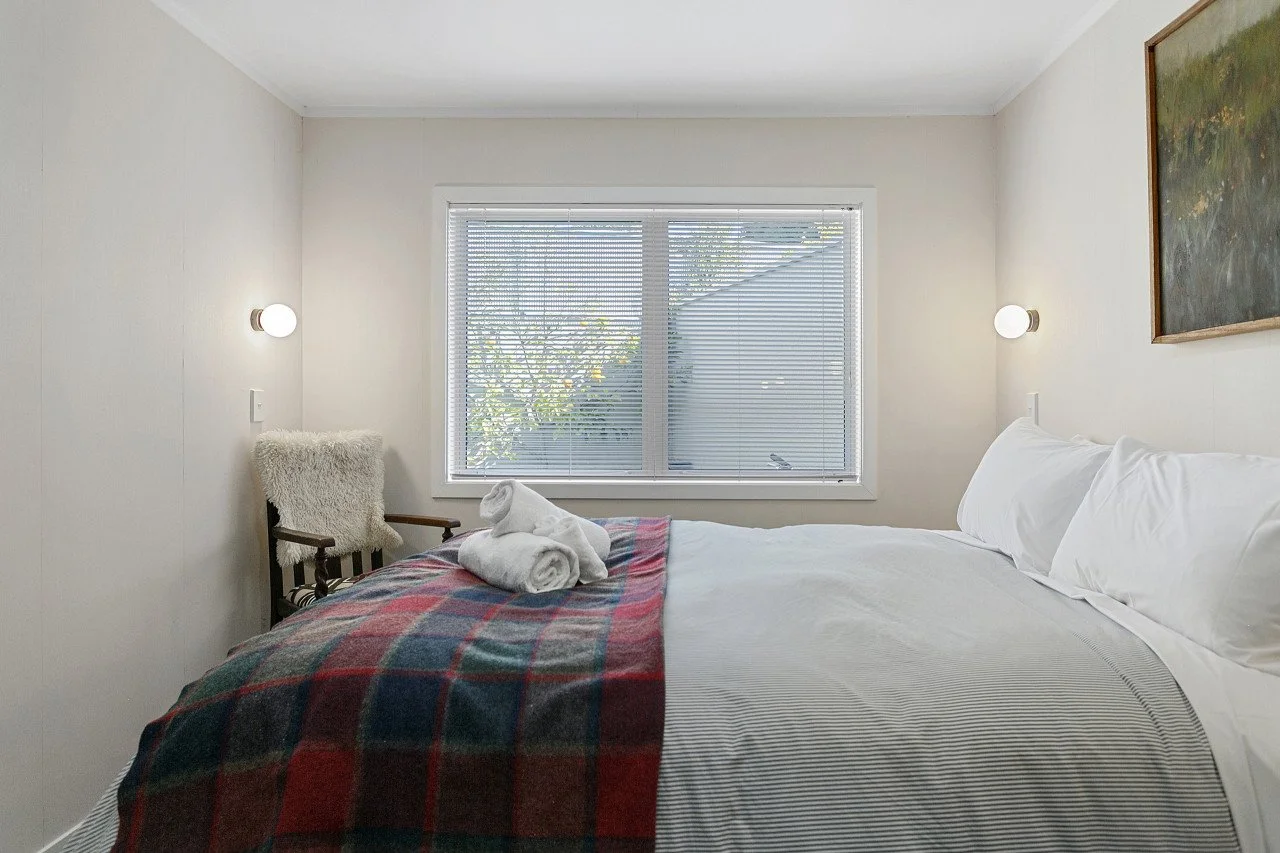 A neatly made bed with white pillows and striped bedsheets, topped with a folded plaid blanket and towel swans, in a bright room with a large window and wall-mounted lamps.
