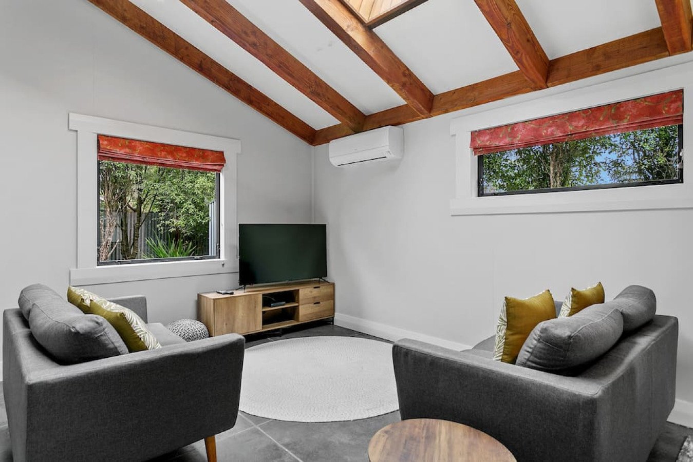 Modern living room with gray sofas, a wooden TV stand with a flat-screen TV, a white area rug, and windows with red floral shades showing greenery outside, wooden ceiling beams, and an air conditioning unit.