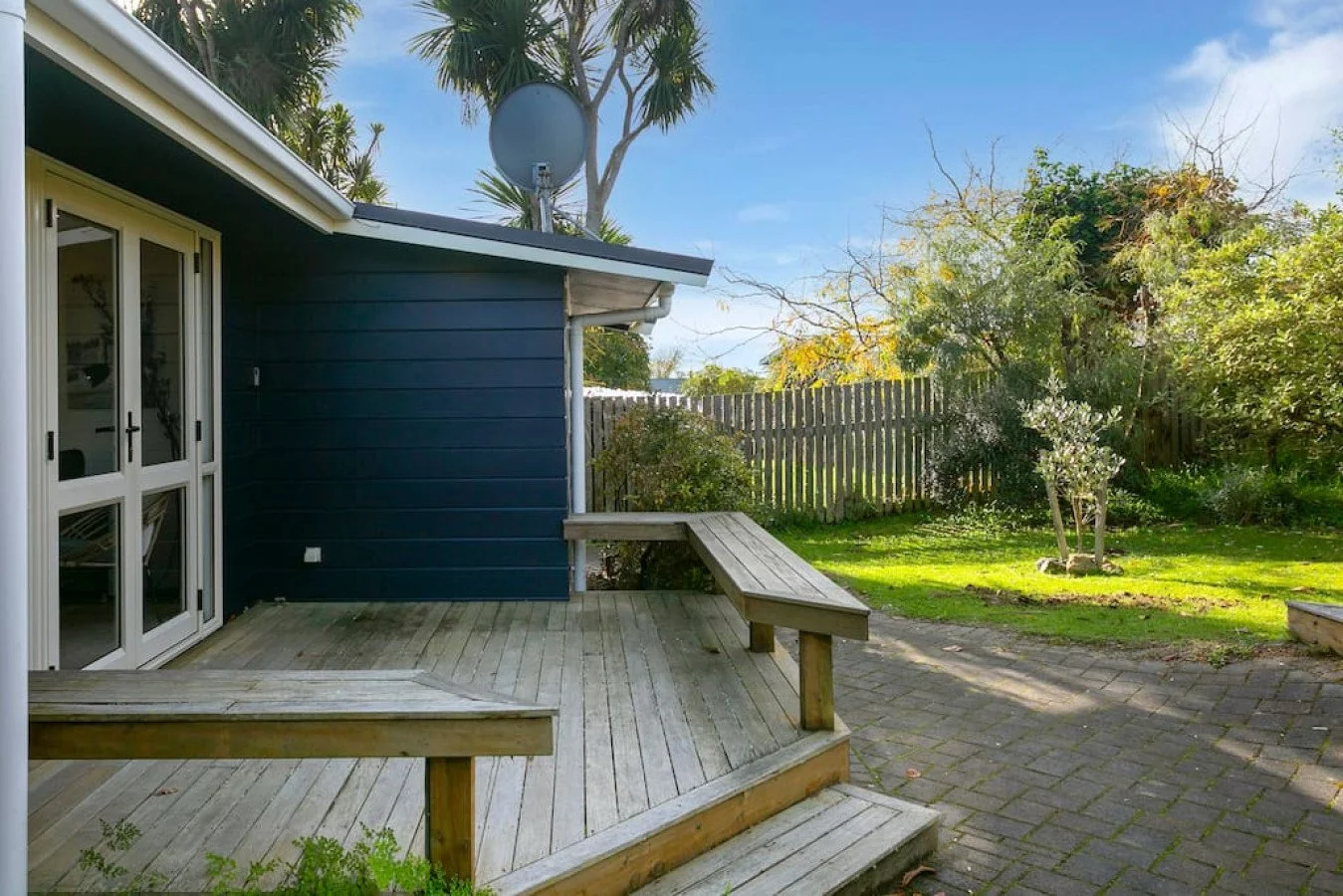 Backyard patio with wooden deck, sliding glass door, blue house wall, green grass, and trees with a wooden fence in the background under a clear sky.