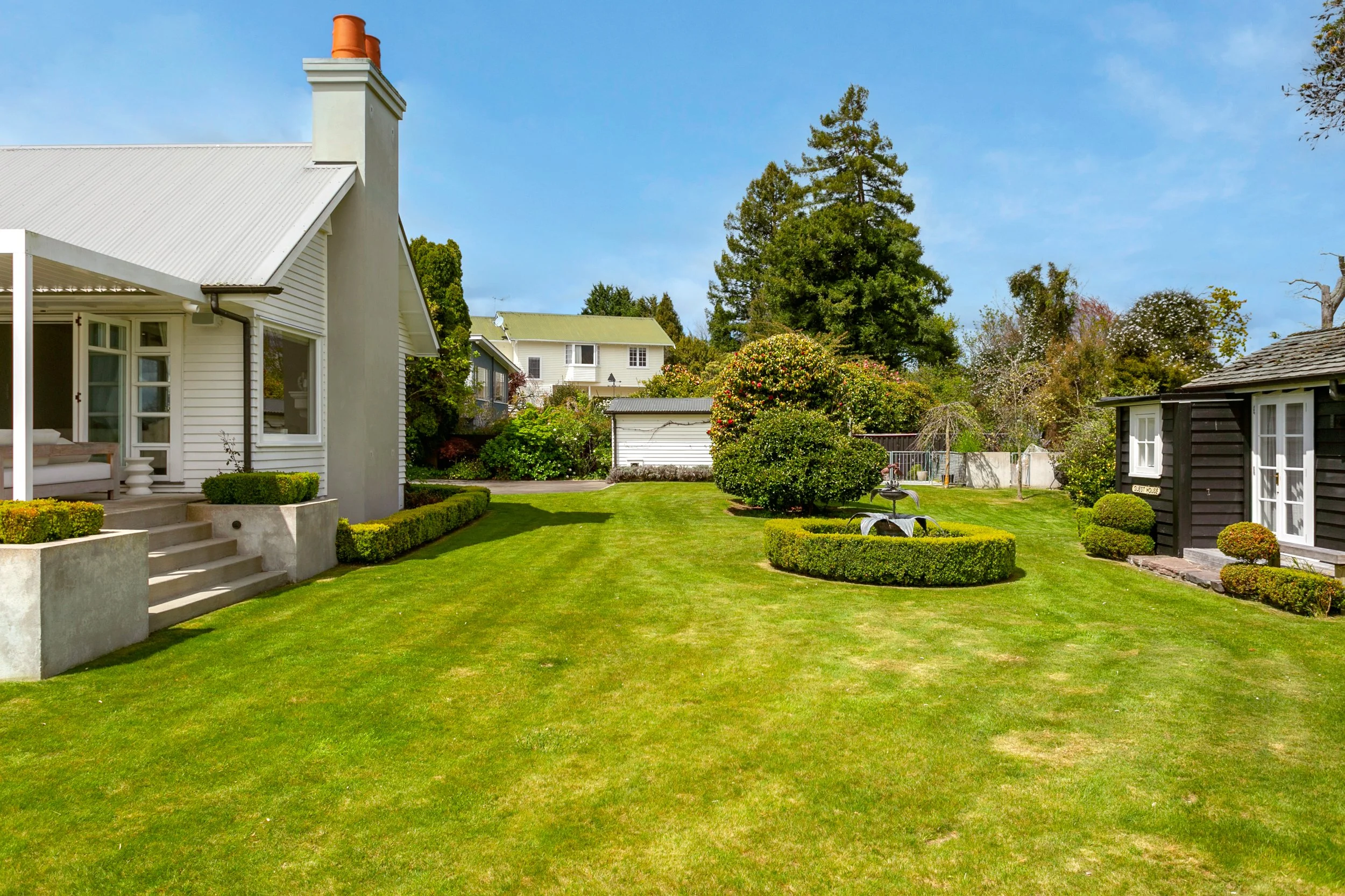 A well-maintained backyard with lush green grass, trimmed bushes, and trees, featuring a white house on the left and a black shed on the right, under a clear blue sky.
