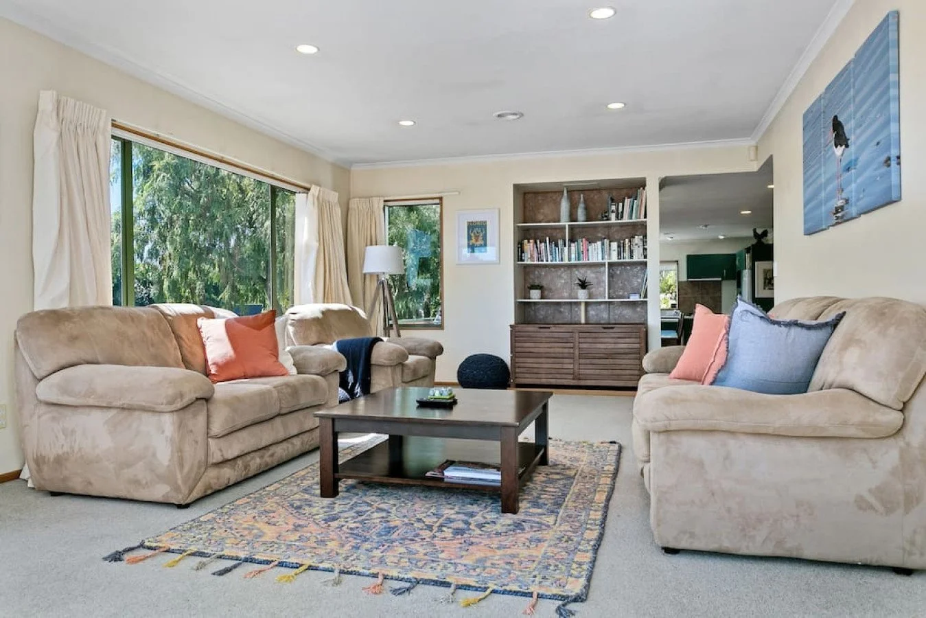 Living room with two beige sofas, a wooden coffee table, a patterned rug, window with cream curtains, bookcase with decor, and wall art of a person on water.