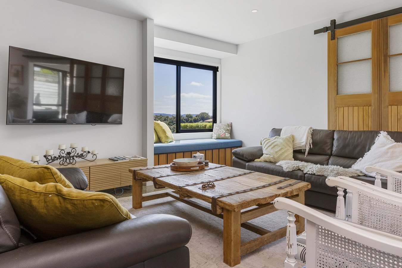 Living room with a wall-mounted TV, a window seat with green and floral pillows, a brown leather sofa with white and cream pillows, a rustic wooden coffee table, a white side chess, and sliding barn doors.