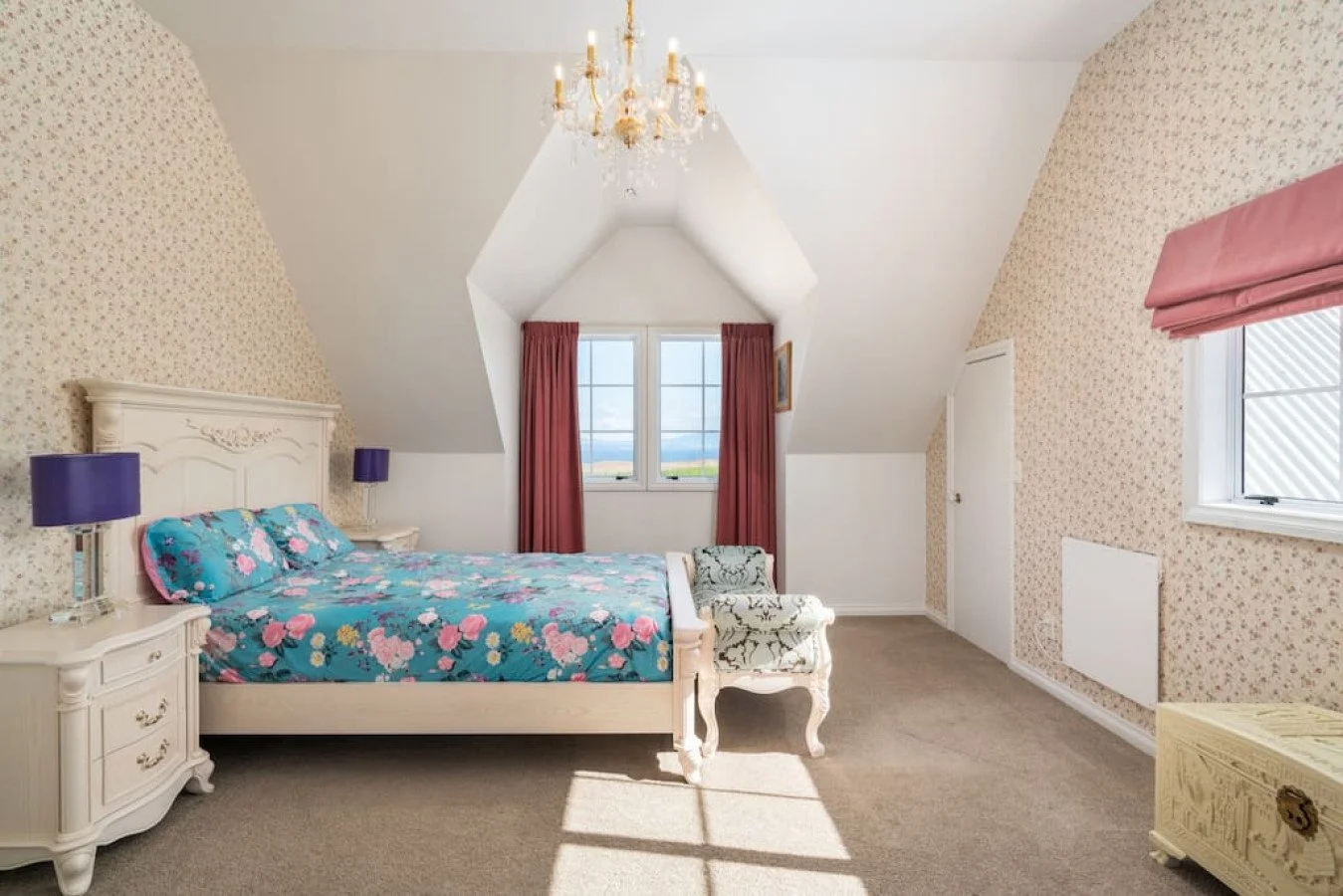 A bedroom with a sloped ceiling, floral wallpaper, a chandelier, a large window with red curtains, a white bed with a floral duvet, two purple lamps on nightstands, a patterned armchair, and a small white cabinet.