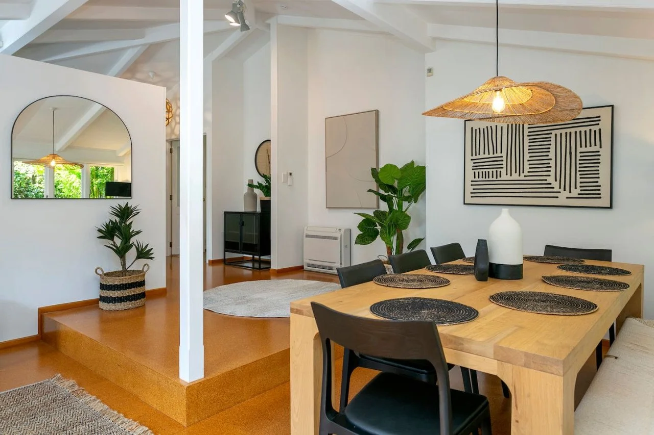 A modern dining area with a wooden table set with black placemats, surrounded by black chairs. A large white vase and a small black vase sit on the table. Above, a woven pendant light hangs. The wall has two abstract art pieces, and there is a large 