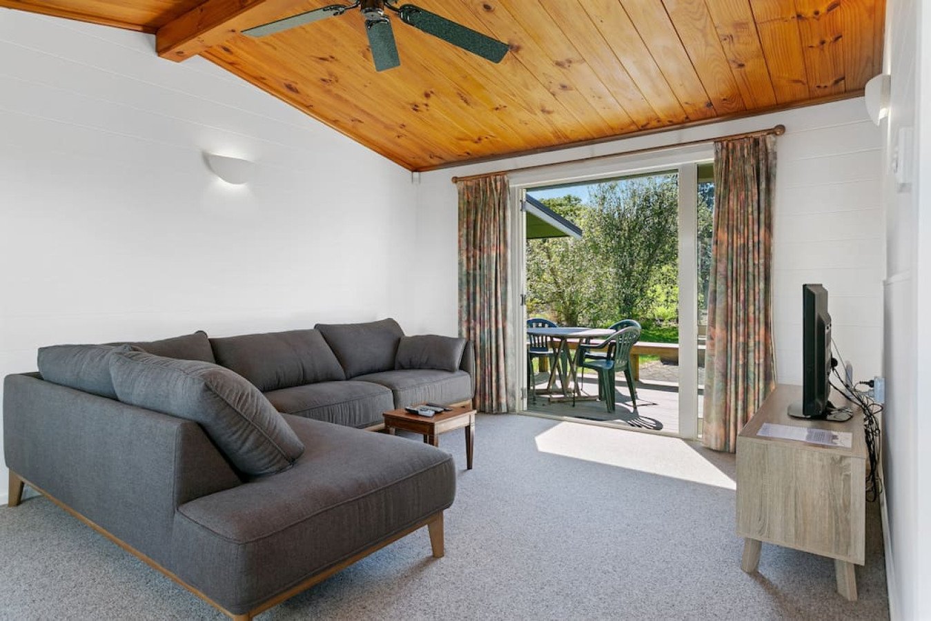 Living room with gray sectional sofa, small wooden coffee table, sliding glass door opening to outdoor patio with table and chairs, wood-paneled ceiling with ceiling fan, white walls, TV on wooden stand, and patterned curtains.