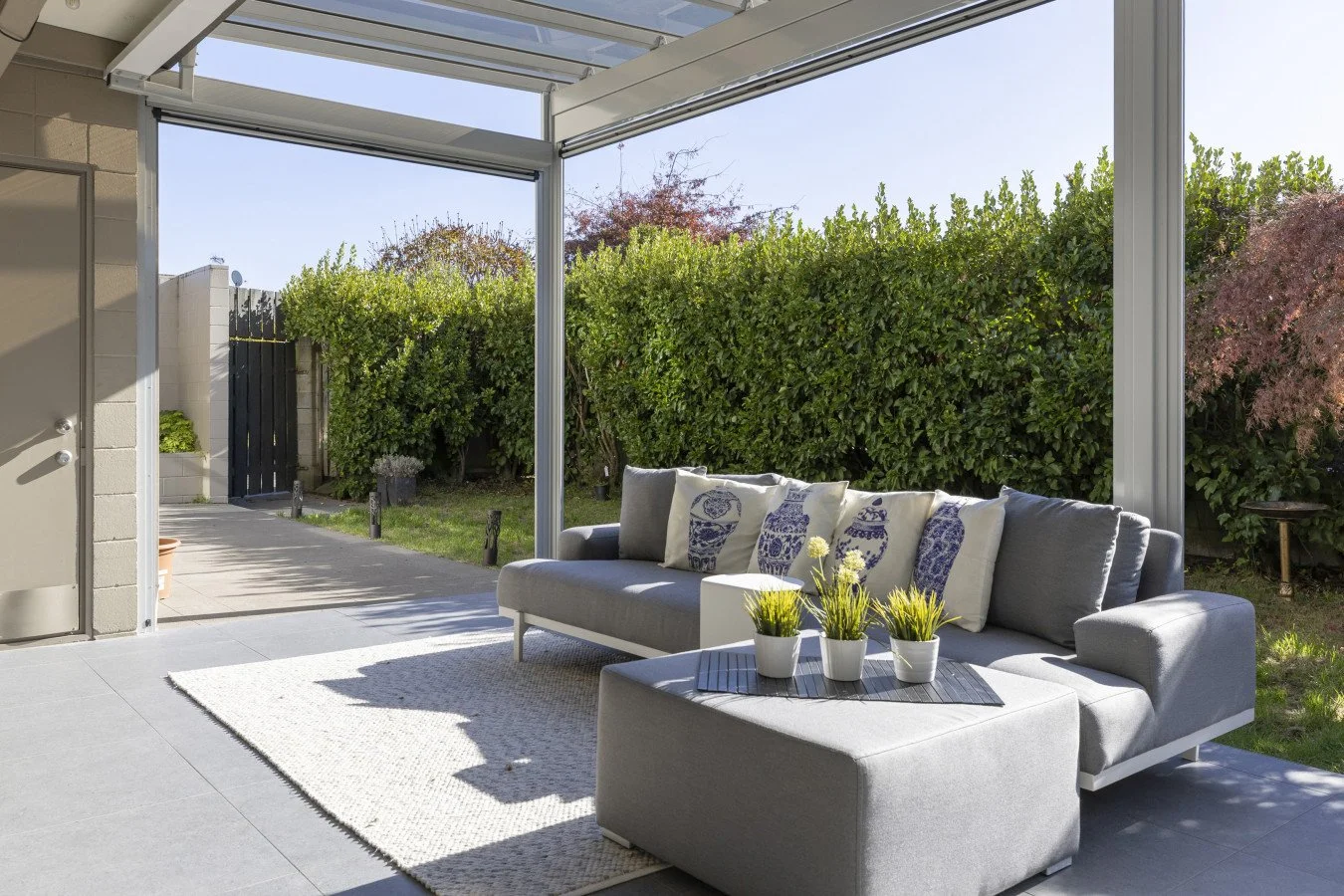 Outdoor patio with a gray sofa and decorative pillows, overlooking a garden with green bushes, sunlight casting shadows, and a tiled floor.