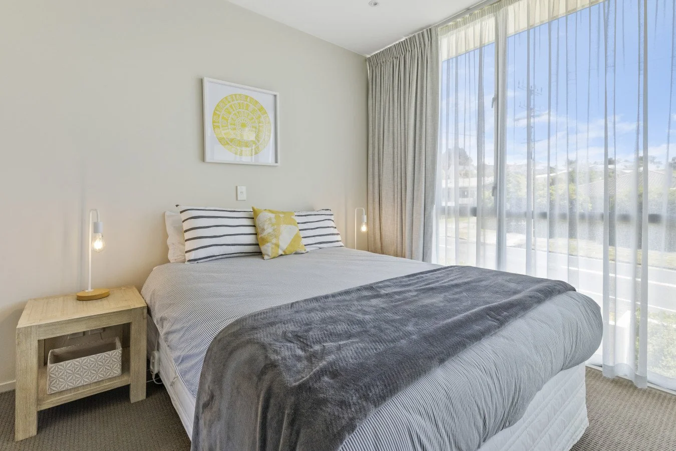 A neatly made bedroom with a bed, decorative pillows, a nightstand with a lamp, a framed art piece on the wall, and large windows with sheer curtains showing a sunny outdoor view.