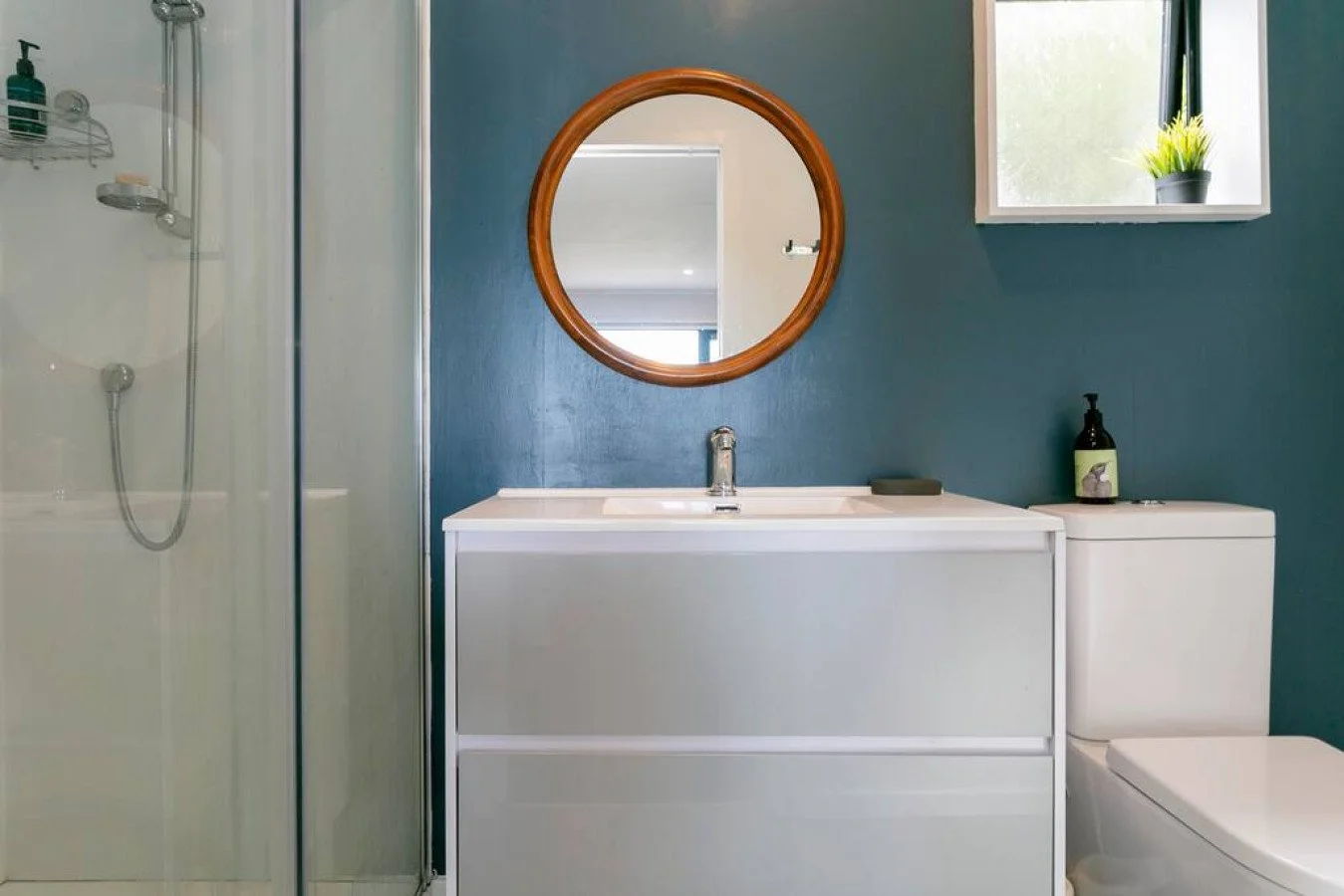 Bathroom with blue wall, round wooden mirror, white vanity with sink, toilet, and small window with a plant on the windowsill.