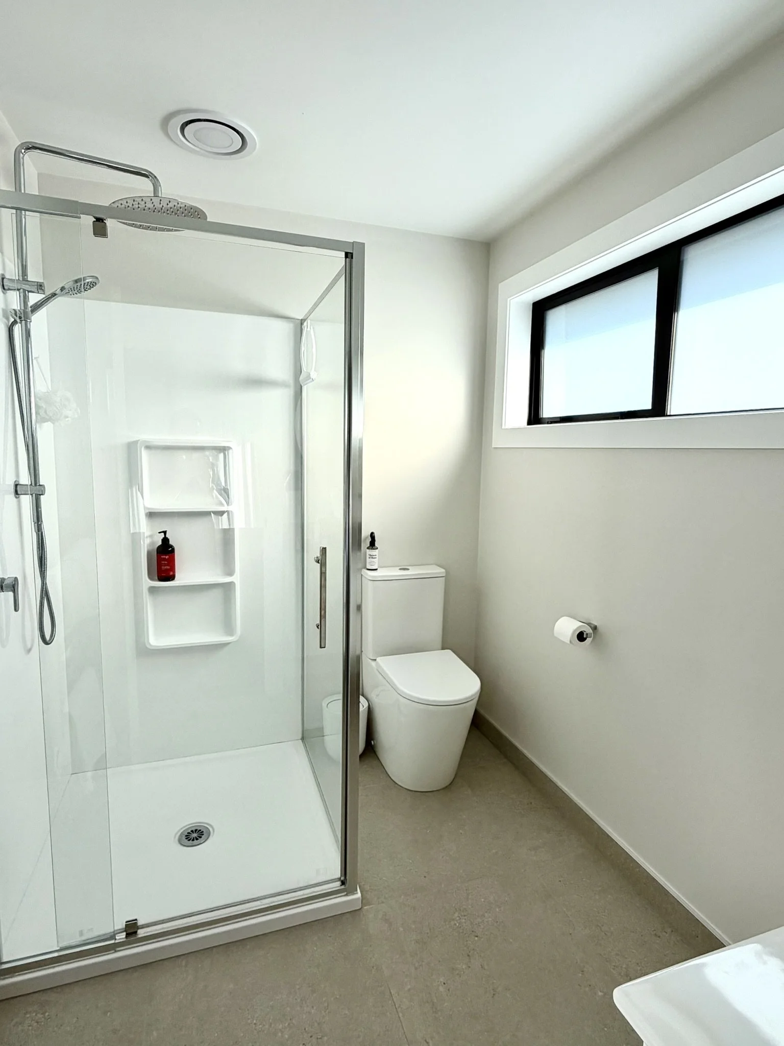 Modern bathroom with a walk-in shower, toilet, and large window, all in white and neutral tones.