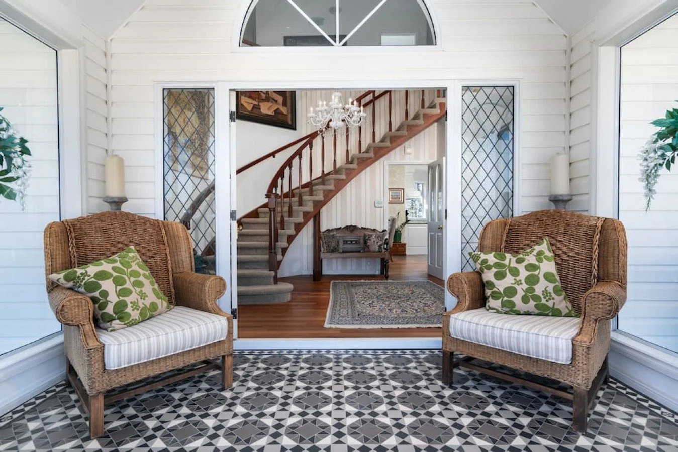 Sunroom with two wicker armchairs, patterned pillows, glass walls, and a view into a living area with a staircase, chandelier, and a small rug.
