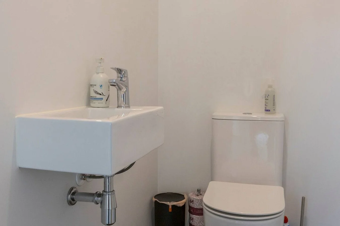 Small bathroom with a white sink, a manual flush toilet, a bottle of hand soap on the sink, and a bottle of disinfectant on the toilet tank.