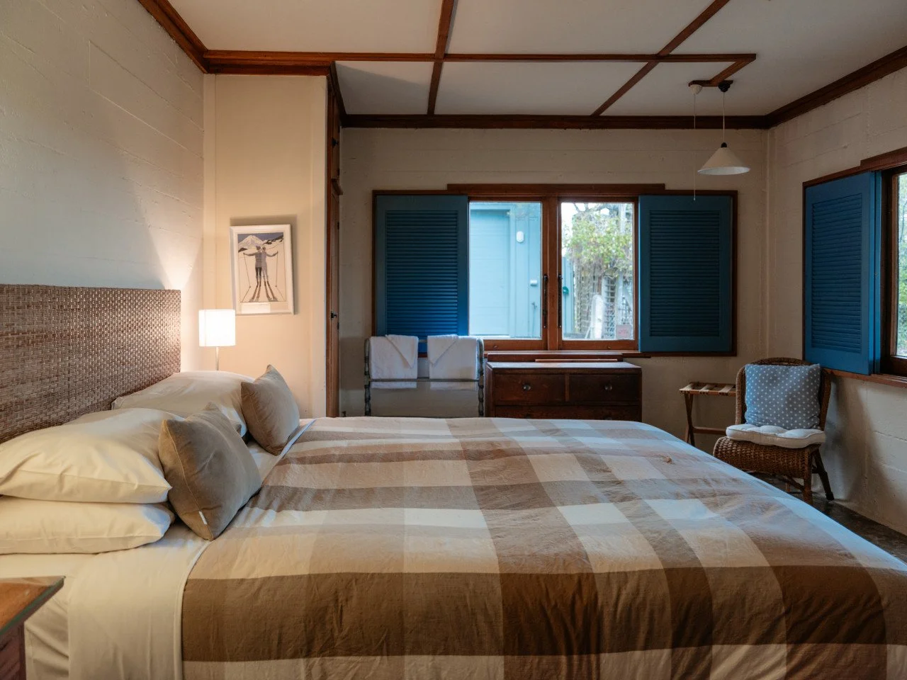 A cozy bedroom with a bed covered in a beige and brown checkered bedspread, pillows, and a headboard. It has a window with blue shutters, a chair with a cushion, and a small dresser beneath the window, with soft lighting from a lamp and natural light