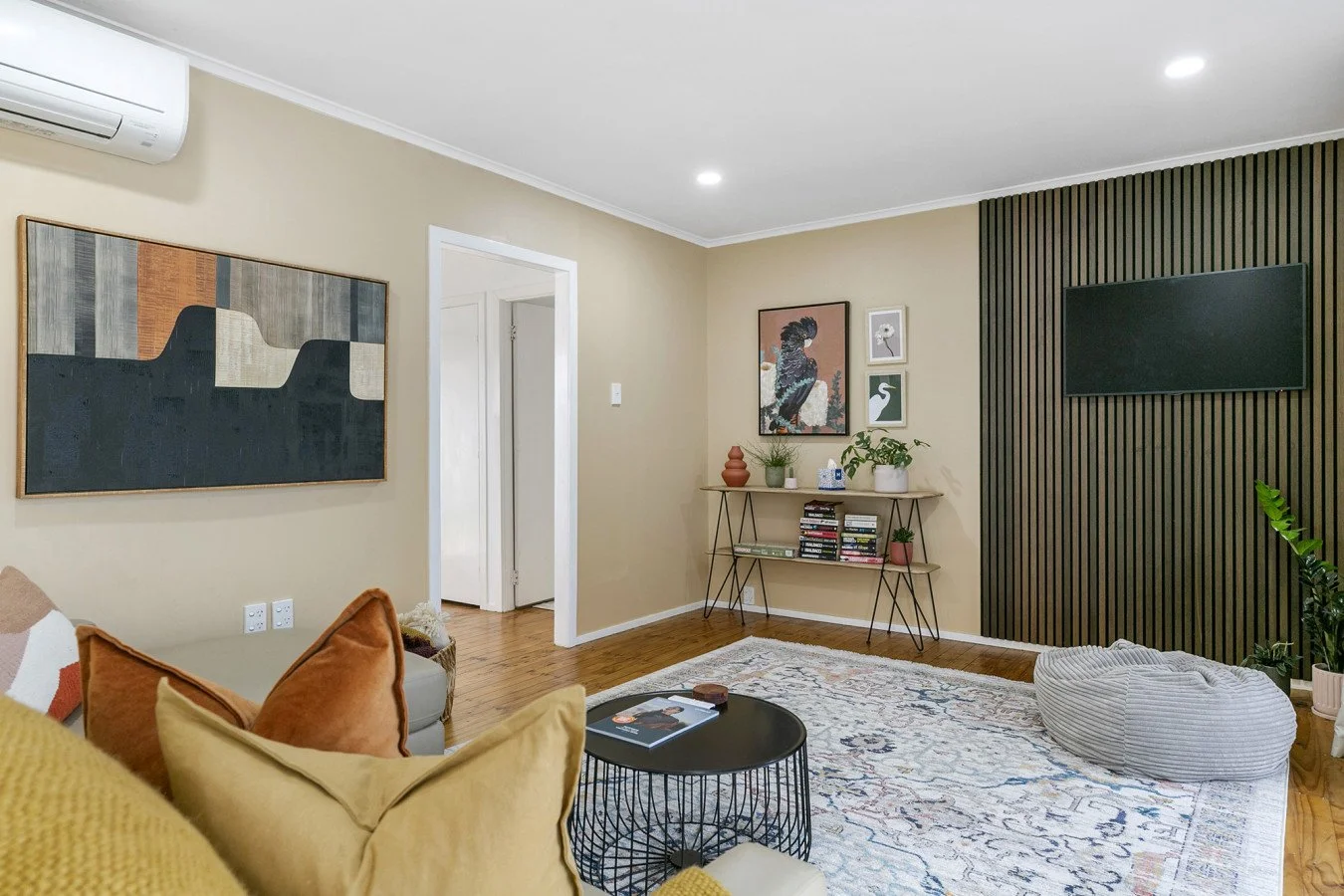 Living room with beige walls, a mounted flat-screen TV, bookshelf, artwork, and colorful cushions on a sofa.
