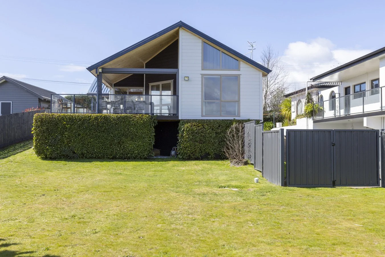 A modern two-story house with white siding and a sloped roof, featuring an upper-level balcony and large windows, surrounded by a well-maintained lawn and a black metal fence.