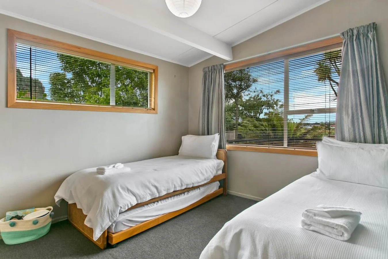 Room with two single beds, white sheets, and towels, large windows with blinds and curtains, view of trees outside.