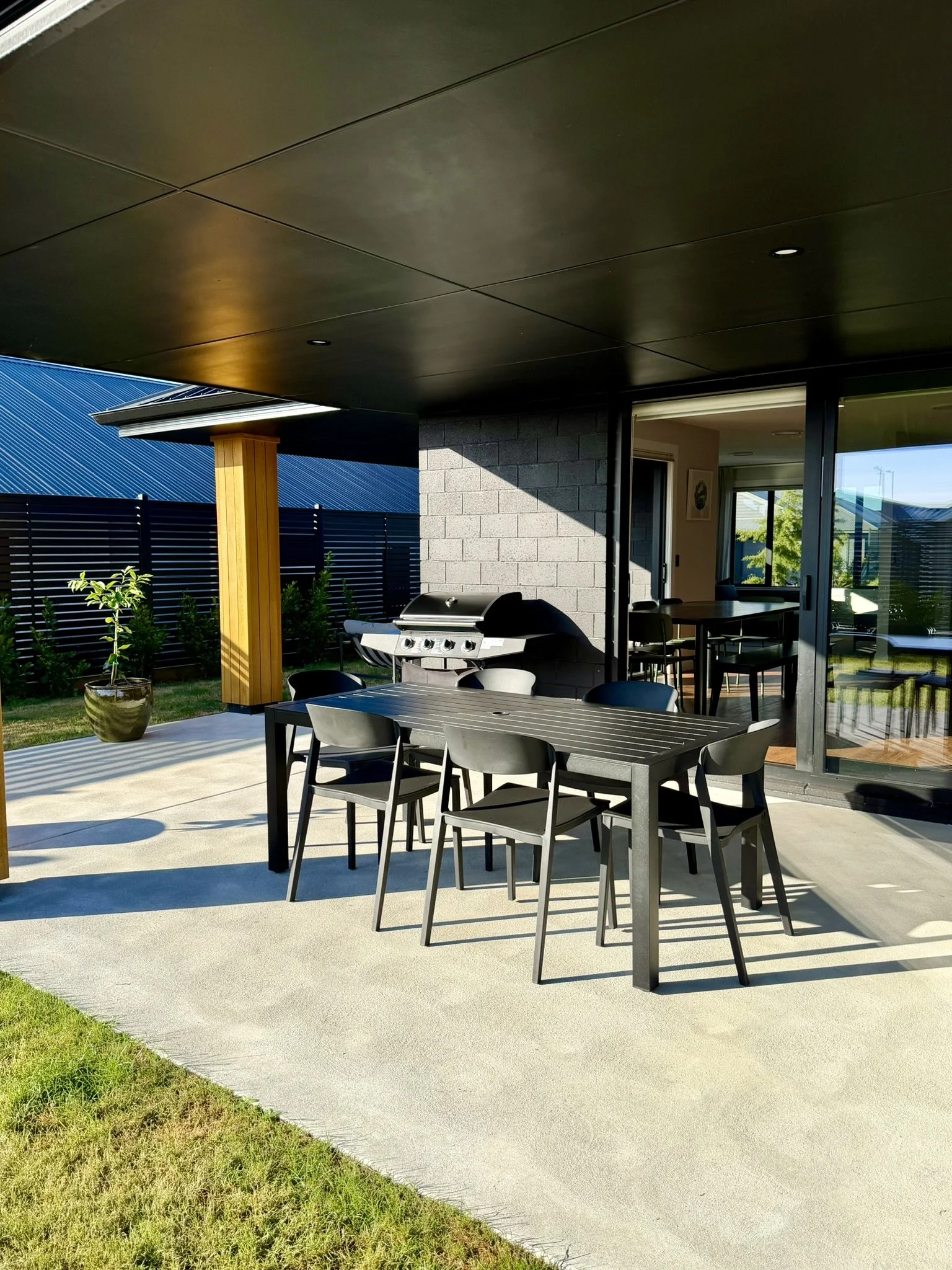 Outdoor patio area with black dining table and chairs, gas grill, potted plant, and a view into the house through sliding glass doors.