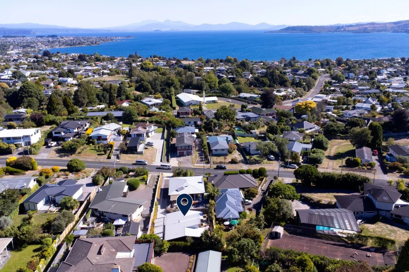 An aerial view of a coastal residential neighborhood with many houses, greenery, and trees, overlooking a large body of water with distant mountains in the background.
