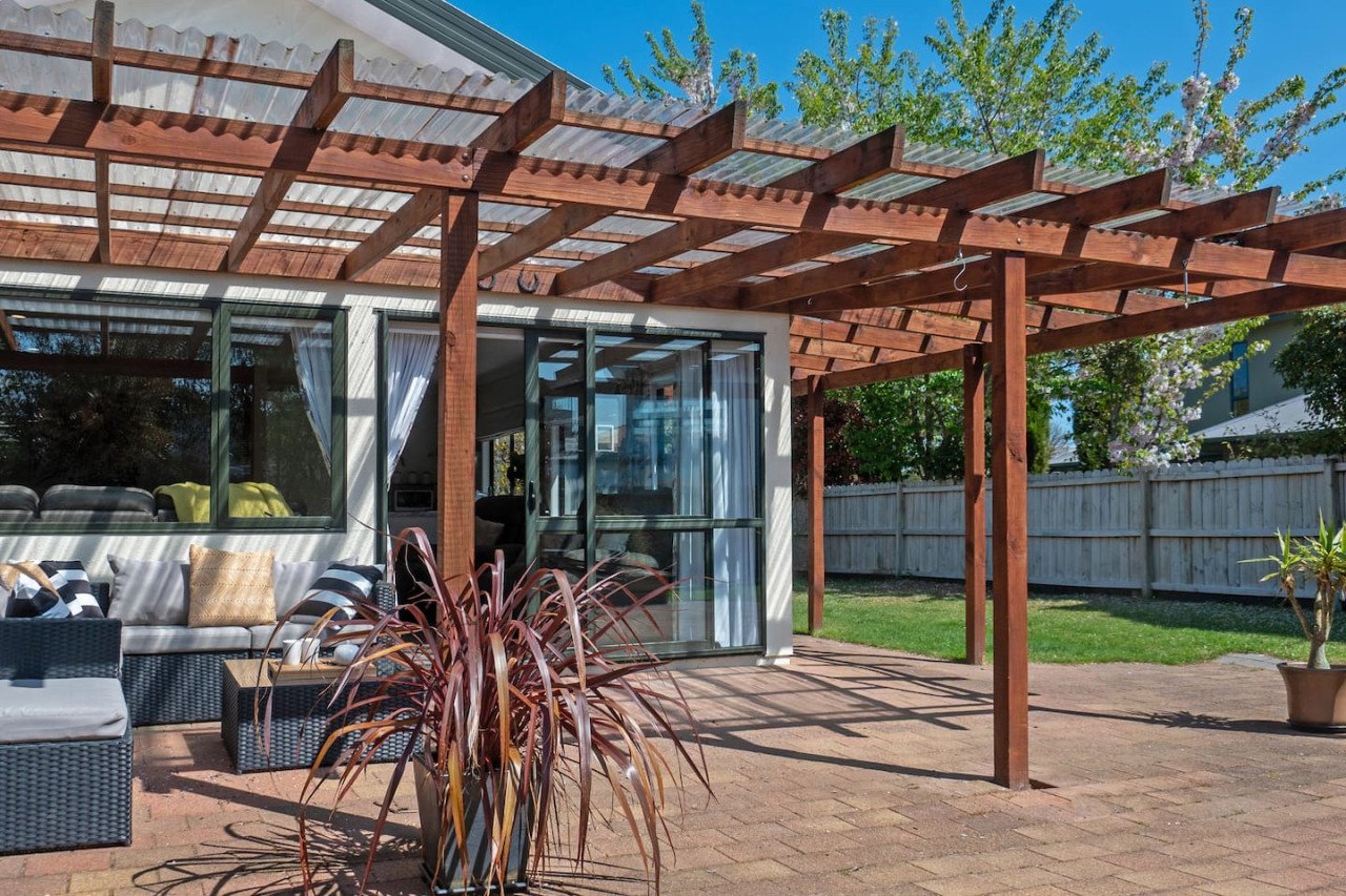 Backyard patio with a wooden pergola, outdoor furniture, potted plants, and a wooden fence, under a clear blue sky.