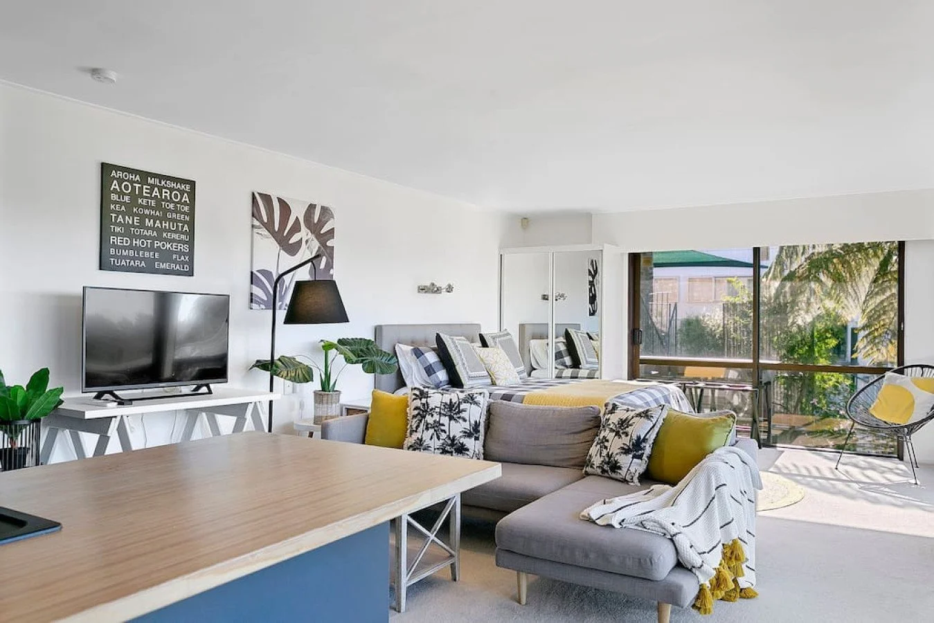 Bright living room with gray sofa, yellow and patterned pillows, a wooden dining table, a TV on a white stand, and large sliding glass doors leading to a balcony with outdoor seating and greenery.