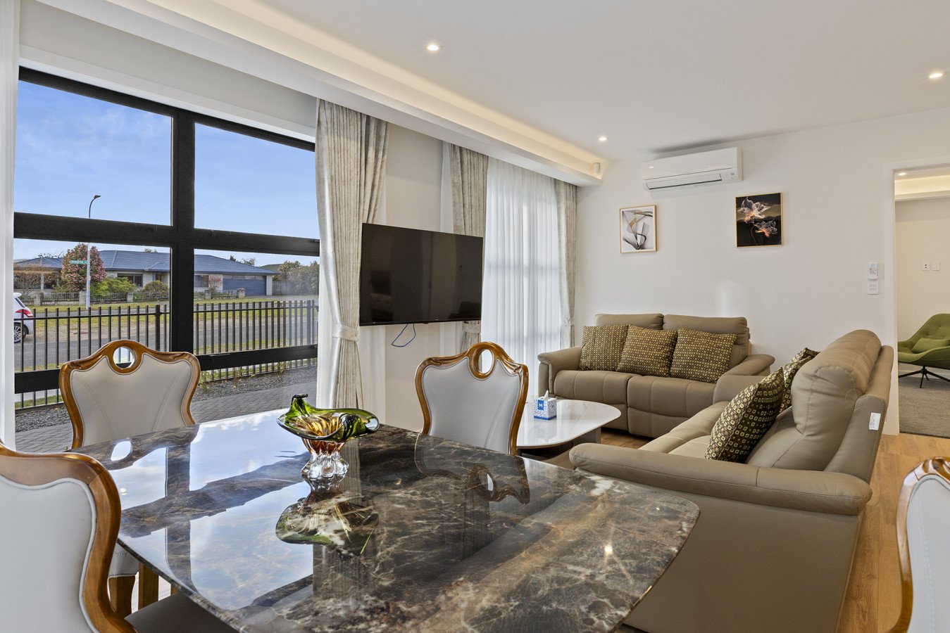 Living room with large window, dining table, beige sofas, wall-mounted TV, wall art, and air conditioning unit.