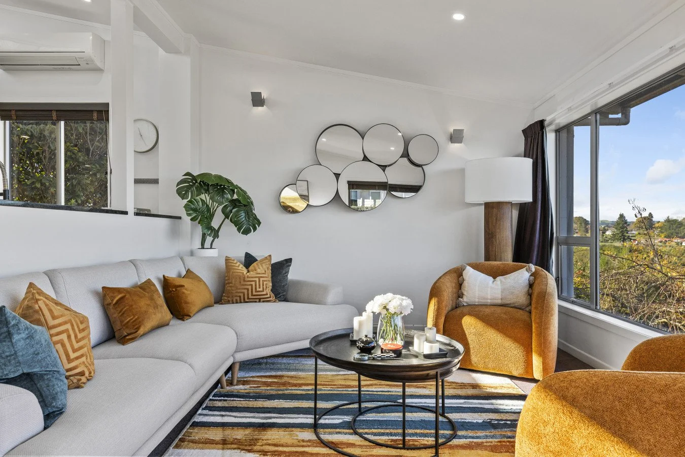 Living room with white sofa, mustard and dark gray pillows, retro style coffee table with candles, large yellow armchair, large window with city view, modern wall mirror art, potted plant, floor lamp, striped rug.