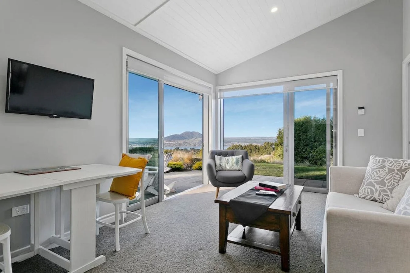 Living room with large windows showing outdoor view, a gray armchair, a white sofa with patterned pillows, a wooden coffee table, and a wall-mounted TV.