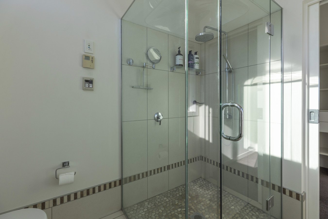 A modern glass shower enclosure in a bathroom with a pebble tile floor and white tiled walls with a decorative border. Inside the shower, a showerhead, a small mirror, and two shampoo bottles are visible.