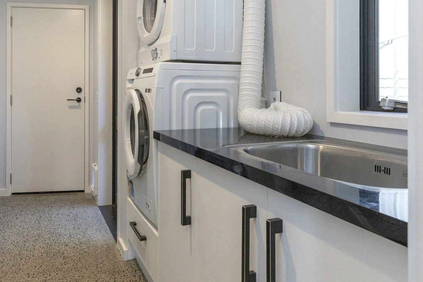 Laundry room with a stacked washer and dryer, a black countertop, a stainless steel sink, a white vent pipe, a window, and a white door.