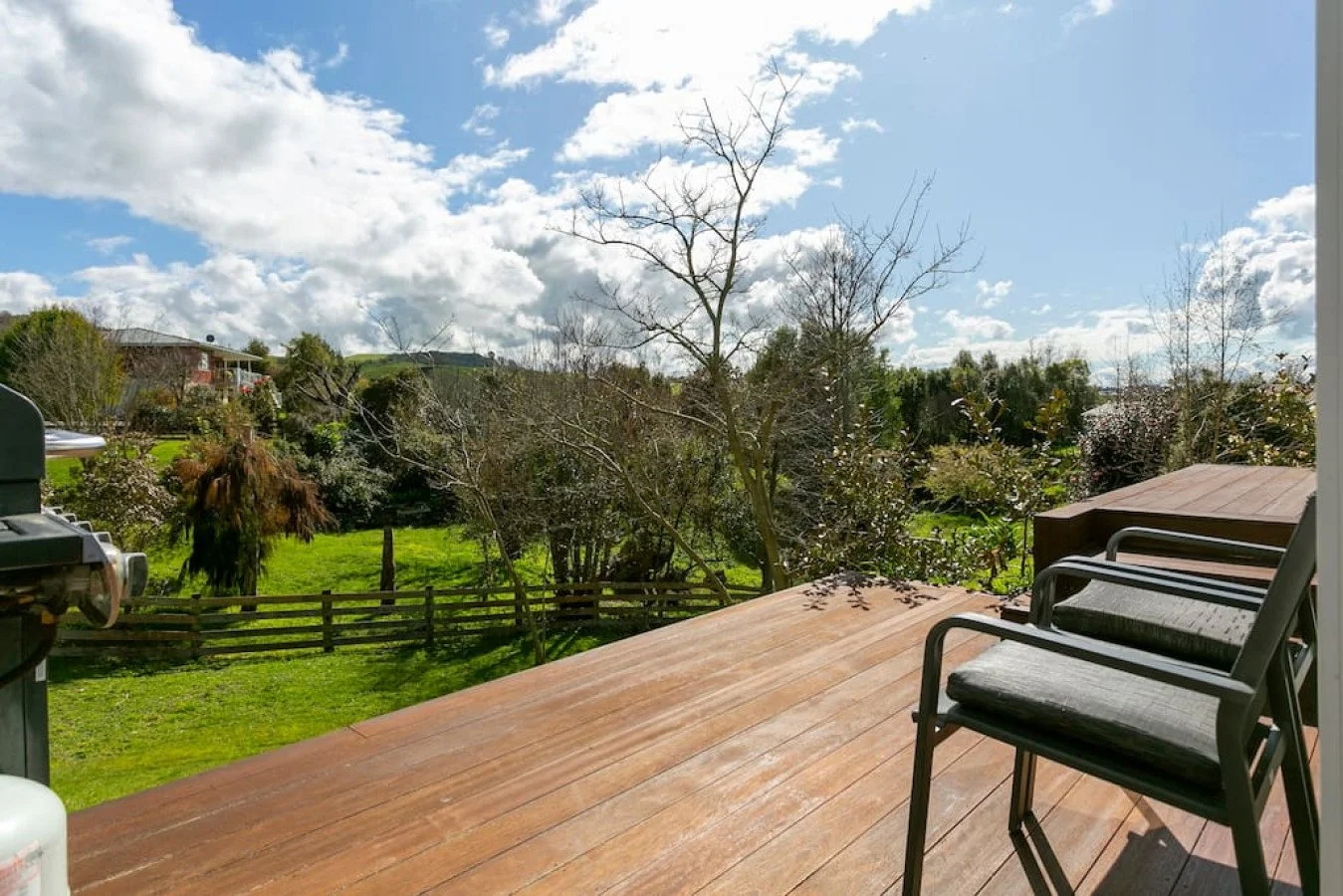 View of a wooden deck overlooking a grassy backyard with trees, a wooden fence, and hills in the distance under a partly cloudy sky.