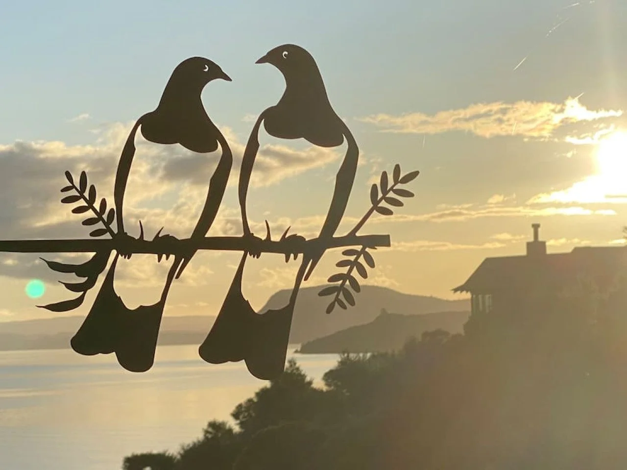 Silhouetted metal artwork of two birds sitting on a branch against a sunset sky with clouds and a distant house on a hill.