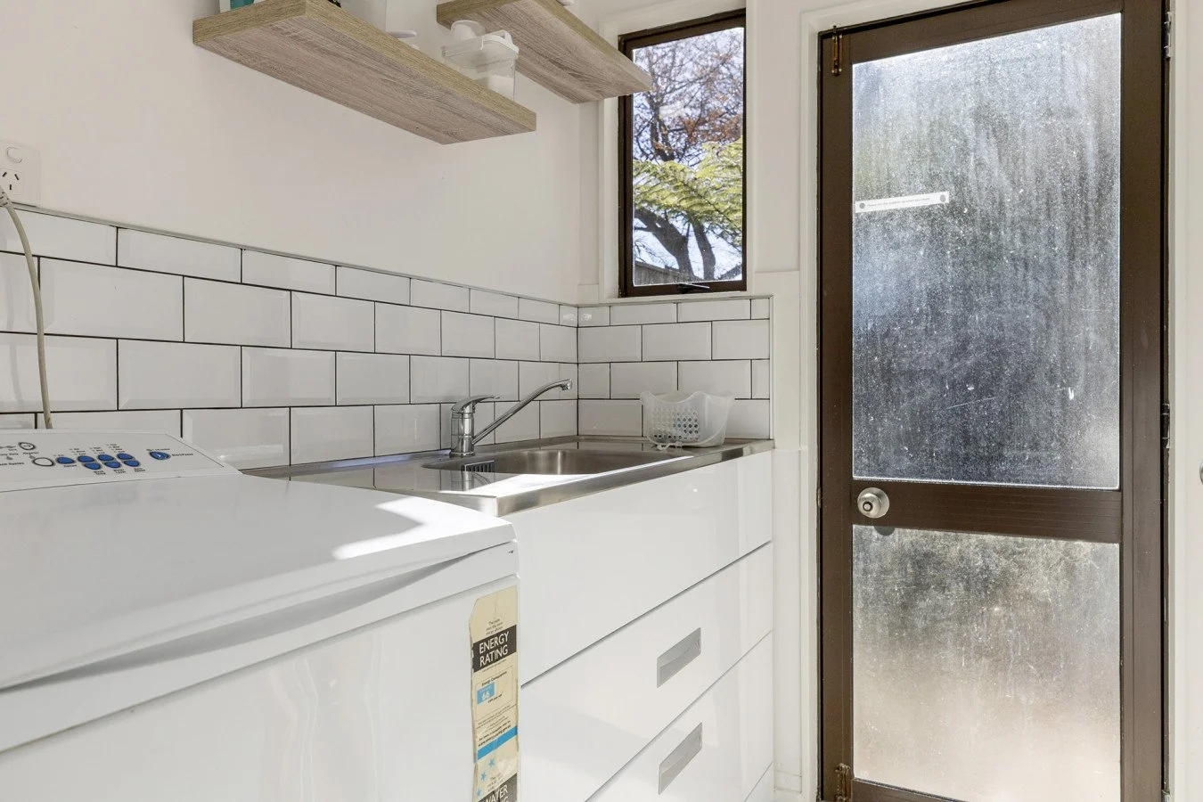 A laundry room with a white washing machine, white cabinetry, a stainless steel sink, white tiled walls, a small window, and a brown door with a glass panel.