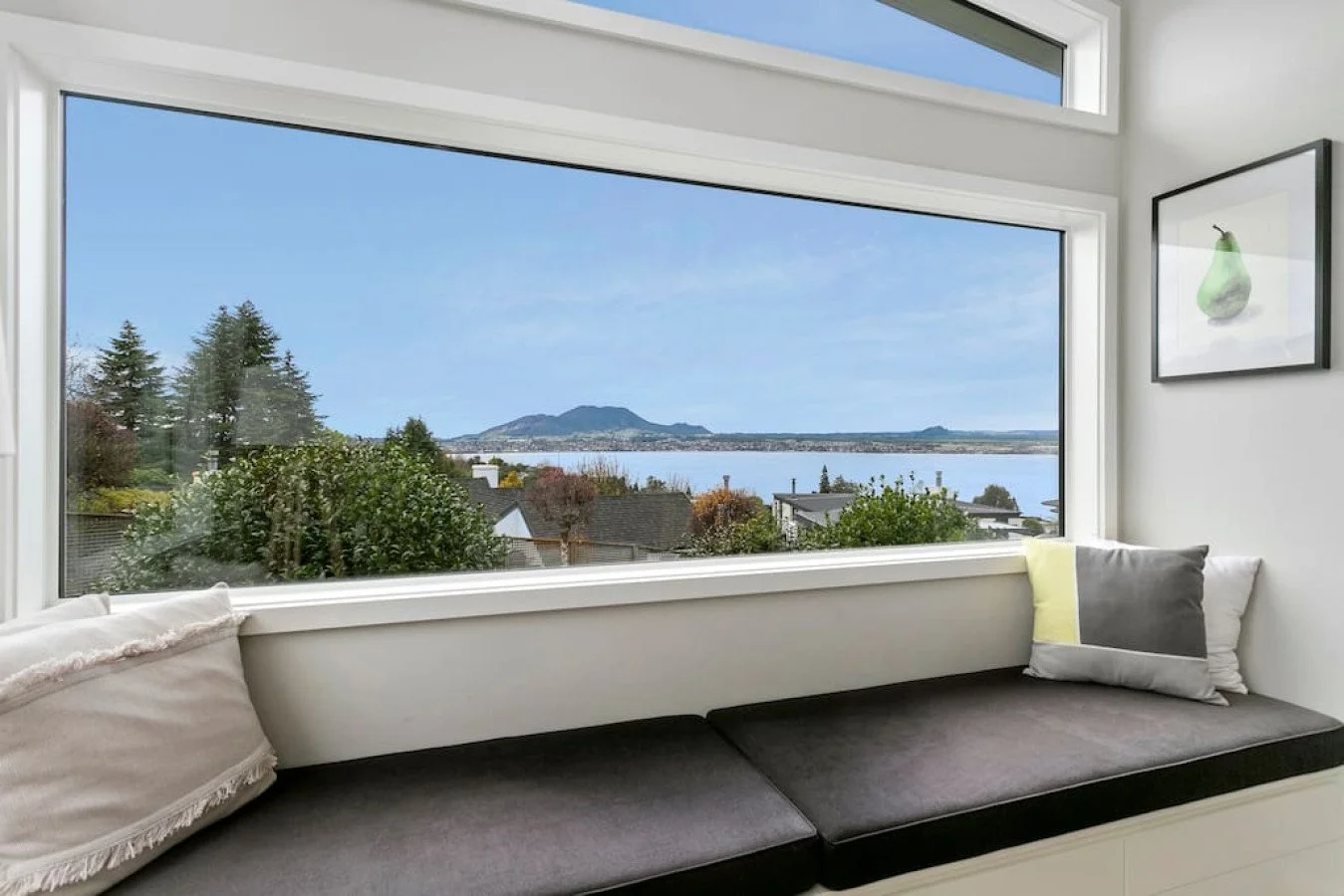 Interior of a modern room with a window seat overlooking a scenic view of trees, houses, a body of water, and mountains in the distance.