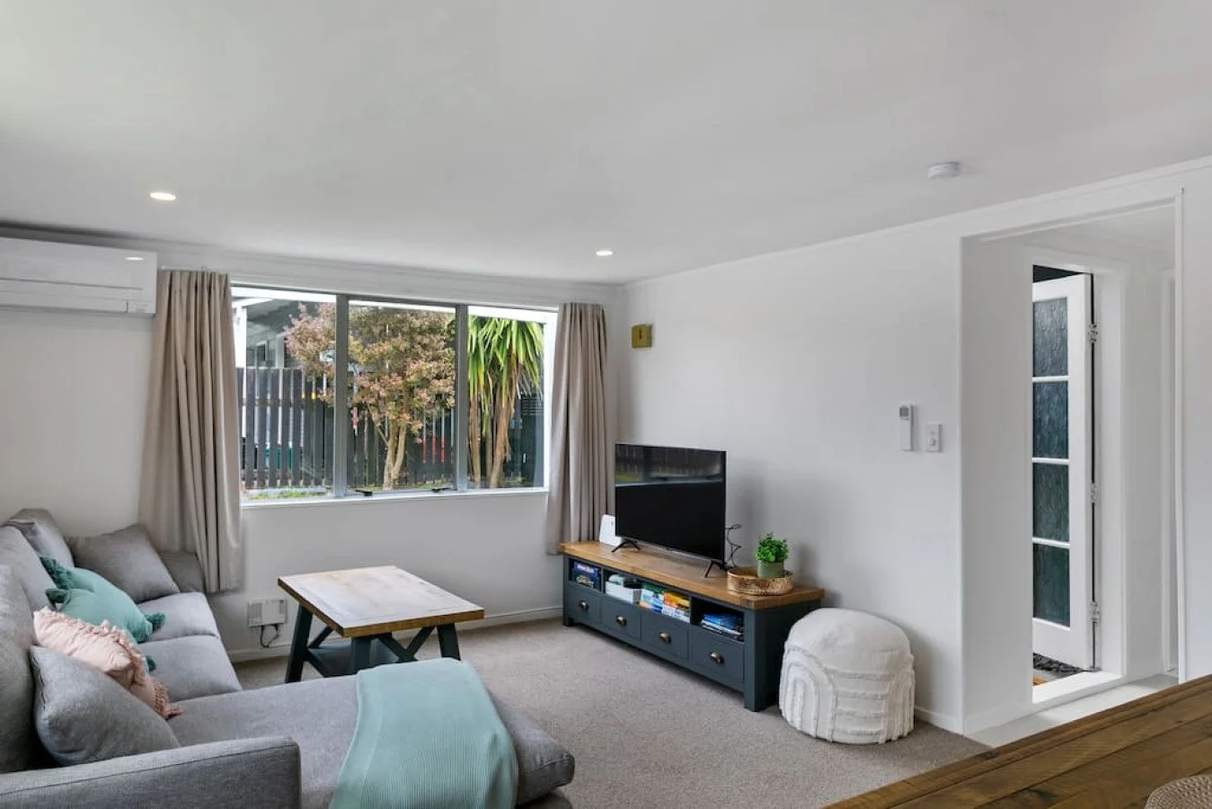 Living room with a gray sofa, a wooden coffee table, a blue TV stand, flat-screen TV, a large window with beige curtains, a potted plant, and an open door leading outside.