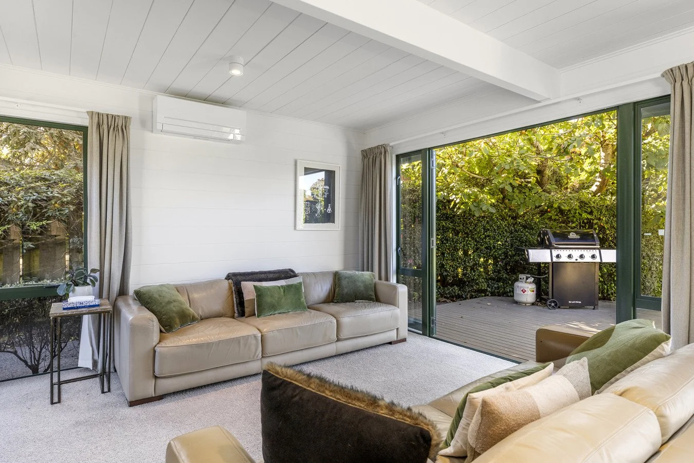 Living room with beige sofas, green and beige cushions, side table with plant and books, large open sliding glass door leading to outdoor deck with grill and greenery.
