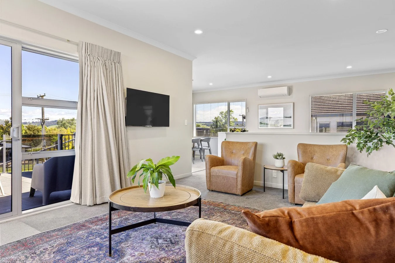Bright living room with beige walls, sliding glass door leading to a balcony, and windows revealing trees and houses outside. Contains brown armchairs, a sofa with cushions, a round coffee table with a potted plant, and a TV on the wall. Decorated wi