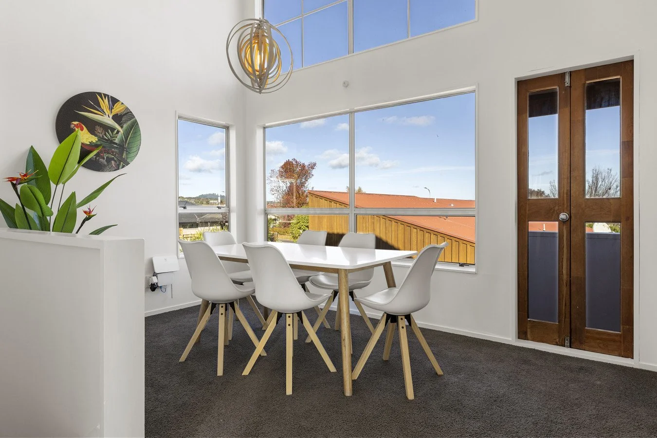 Interior of a bright dining room with a white table and six white chairs, large windows, a wooden door, and decor including a plant and wall art.