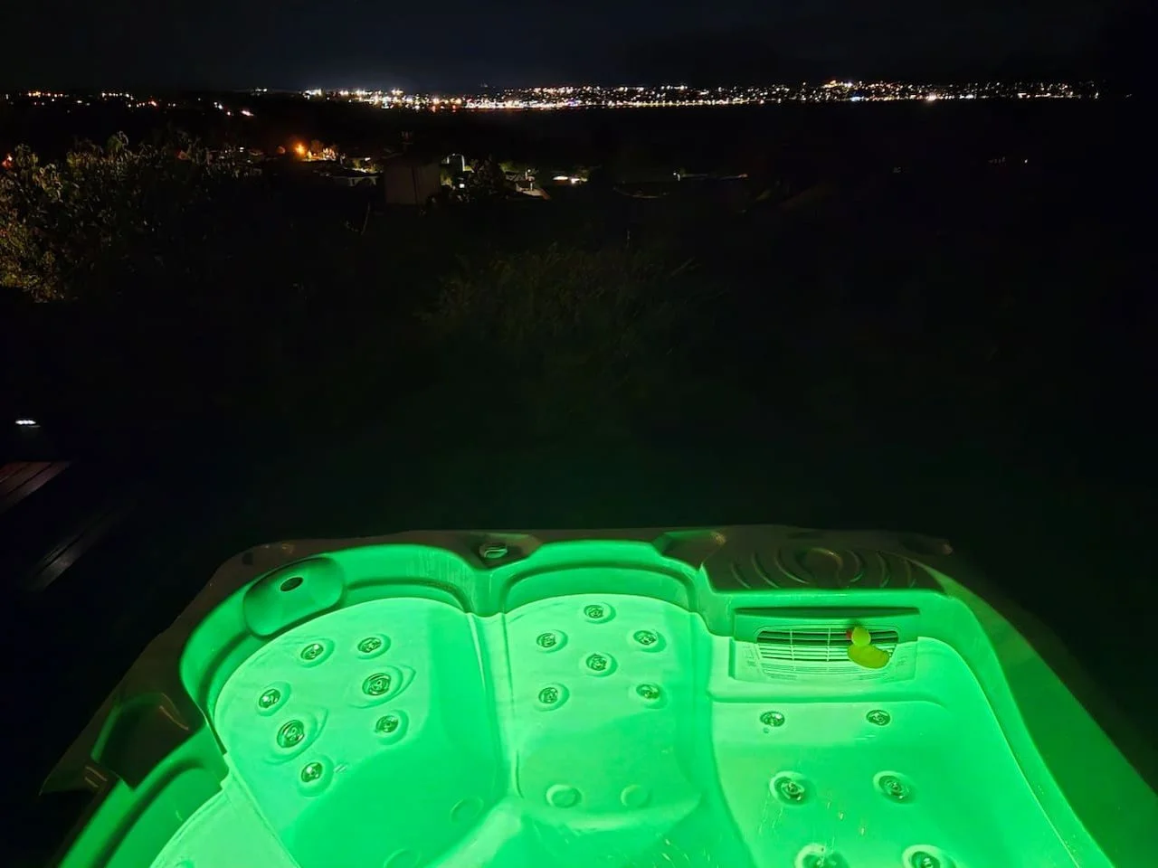 Nighttime view of a glowing green-hot tub overlooking a city skyline with many lights in the distance.