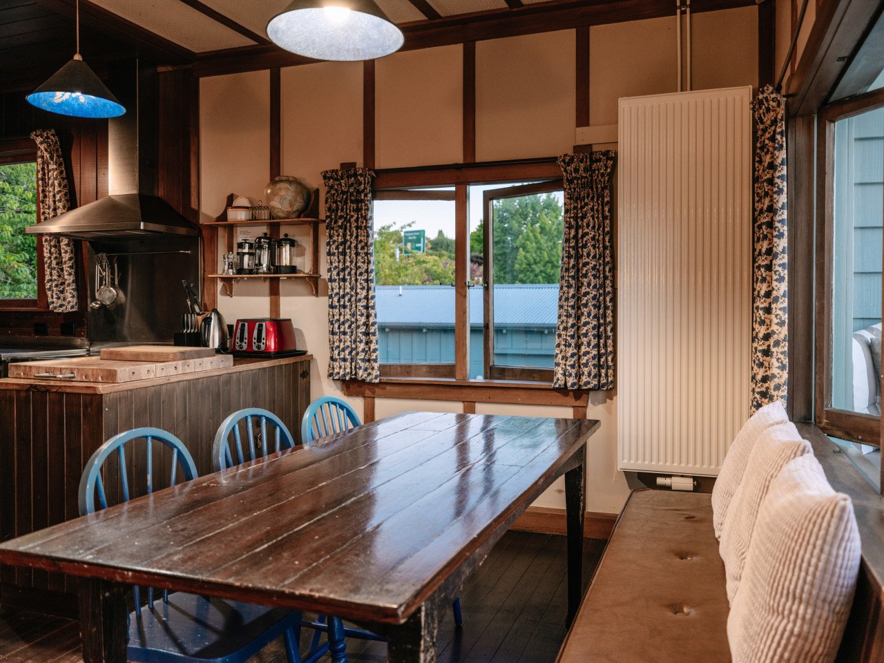 A rustic dining area with a wooden table and blue chairs near windows with floral curtains, overlooking a backyard with a gray fence and trees.