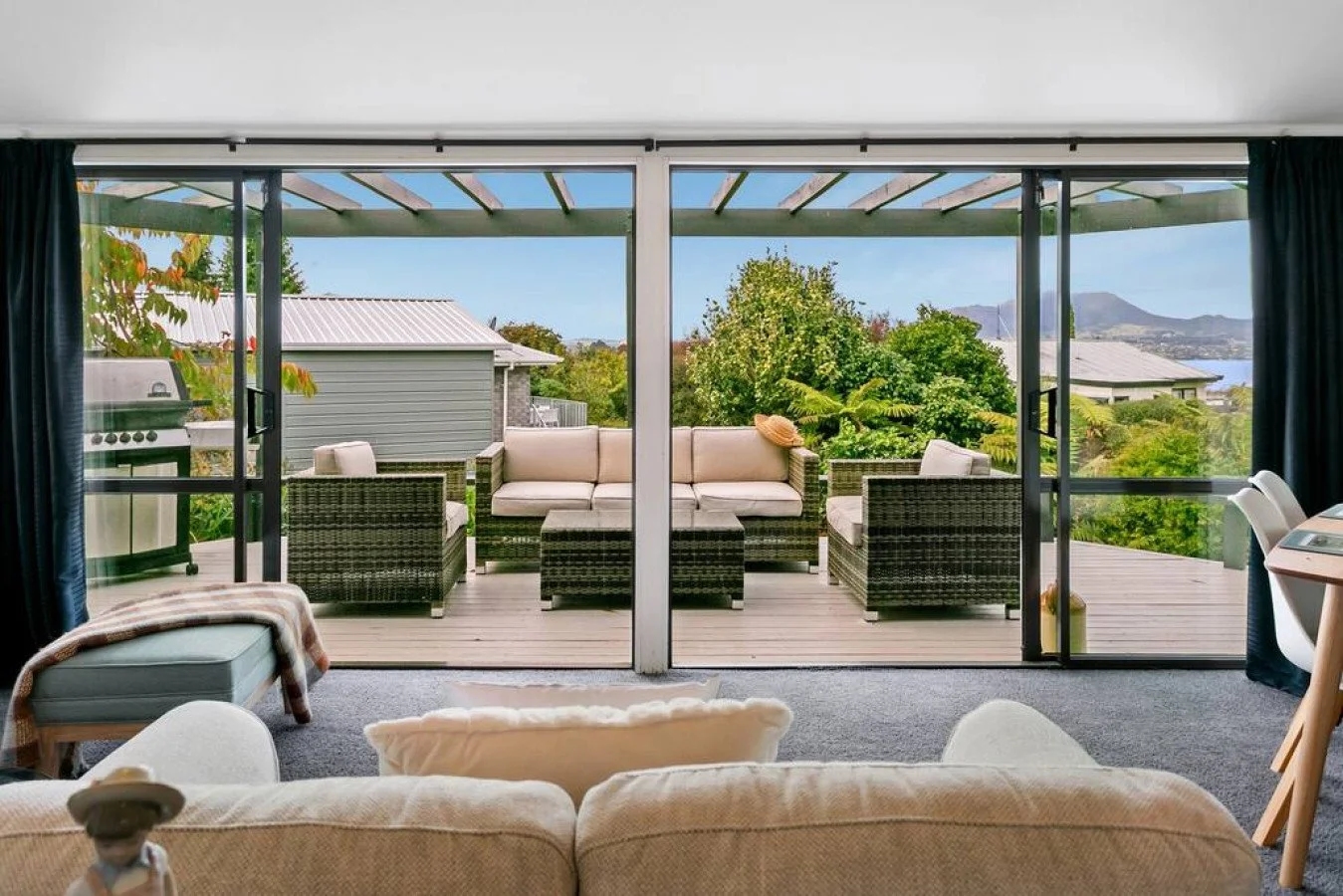 Living room with sliding glass doors opening to a deck with outdoor furniture, lush green trees, a house, and mountains in the distance.