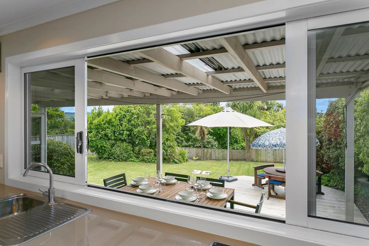 View of a backyard patio seen through a large sliding window from inside a kitchen, with a dining table set outdoors under umbrellas and surrounded by trees and greenery.