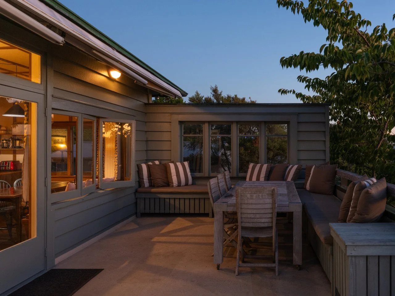An outdoor deck area with a wooden table, chairs, and a corner bench with cushions, adjacent to a house with lit interior lights seen through windows, at dusk.