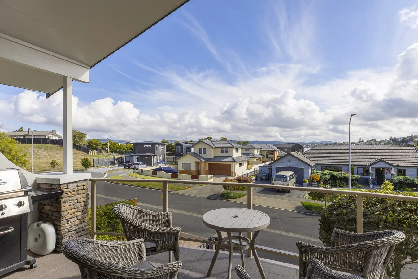 View from a balcony overlooking a suburban neighborhood with houses, lawns, and cars, under a partly cloudy sky.