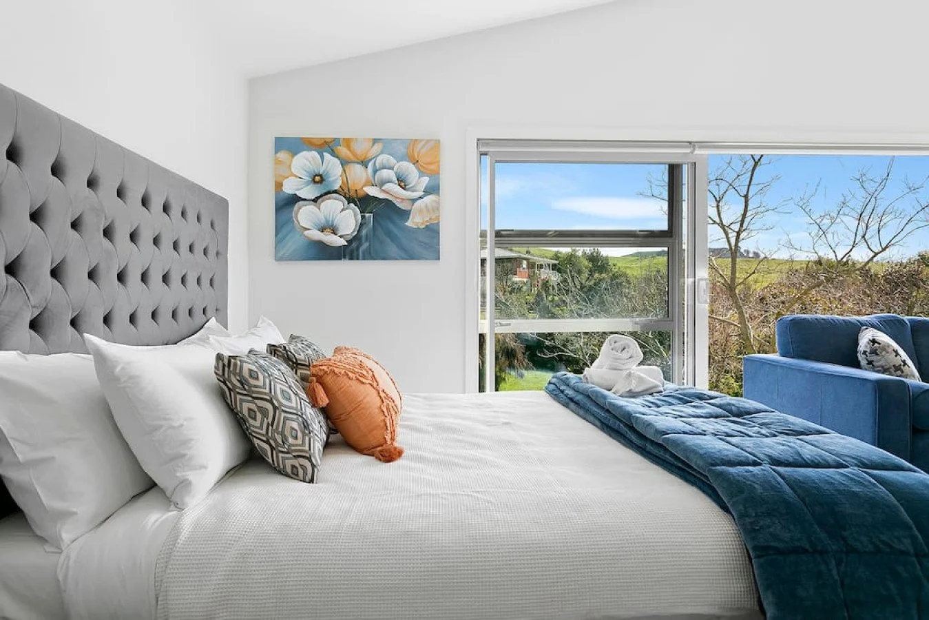A modern bedroom with a large bed featuring gray tufted headboard, white bedding, and decorative pillows. There is a painting of flowers on the wall, and a window showing a scenic outdoor landscape with trees and clear blue sky. A blue armchair with 