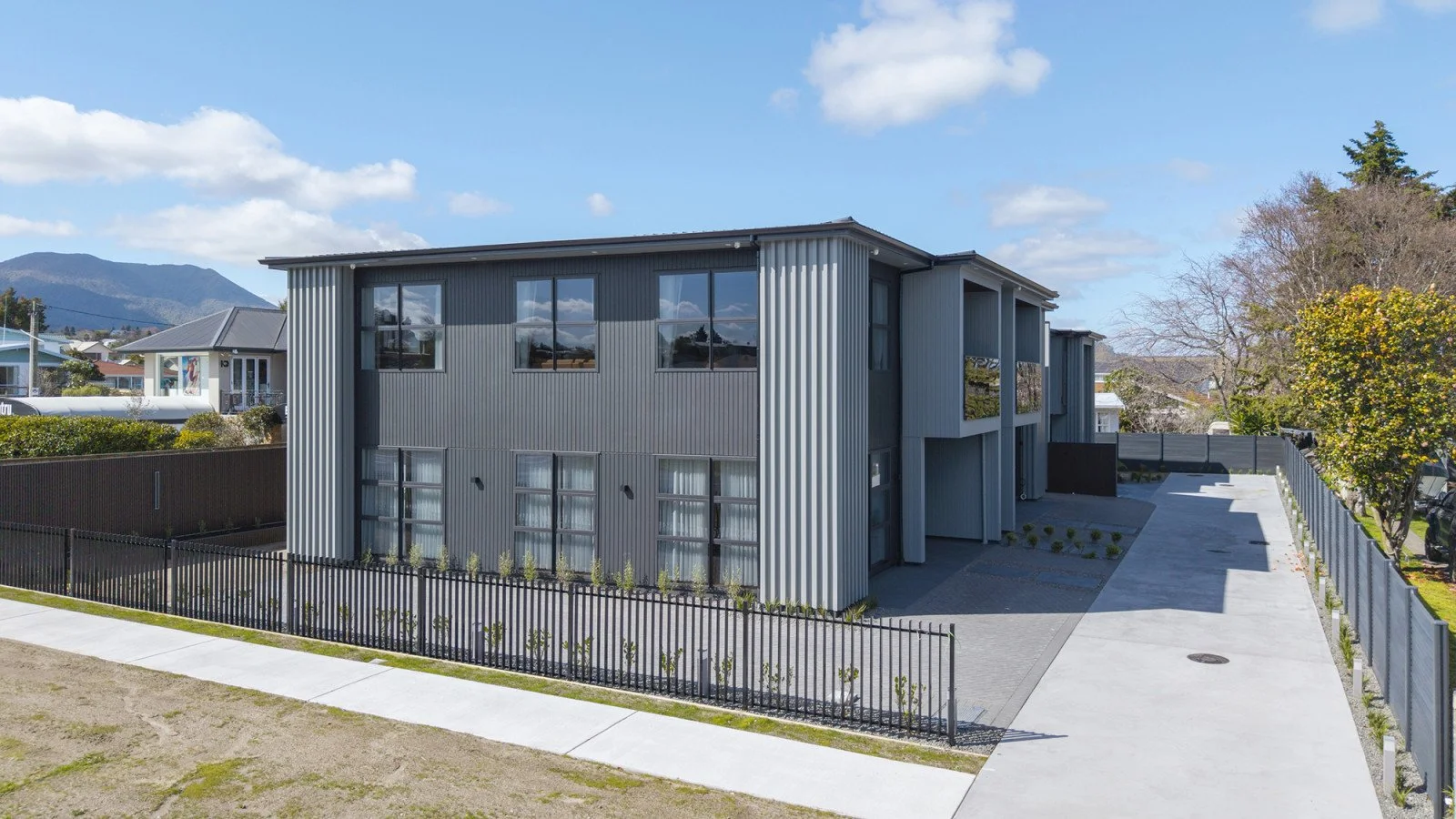 Modern two-story house with gray exterior, metal siding, large windows, and a concrete driveway, surrounded by a black metal fence and some small plants, with cloudy sky and mountains in the background.