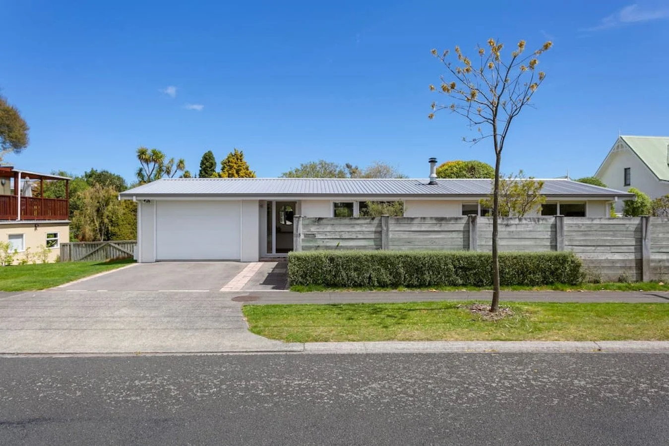A modern single-story house with a white exterior, gray roof, and a driveway with a parked car. The house has a small front yard with a tree and neatly trimmed bushes, and is enclosed by a wooden fence. There are trees and neighboring houses in the b