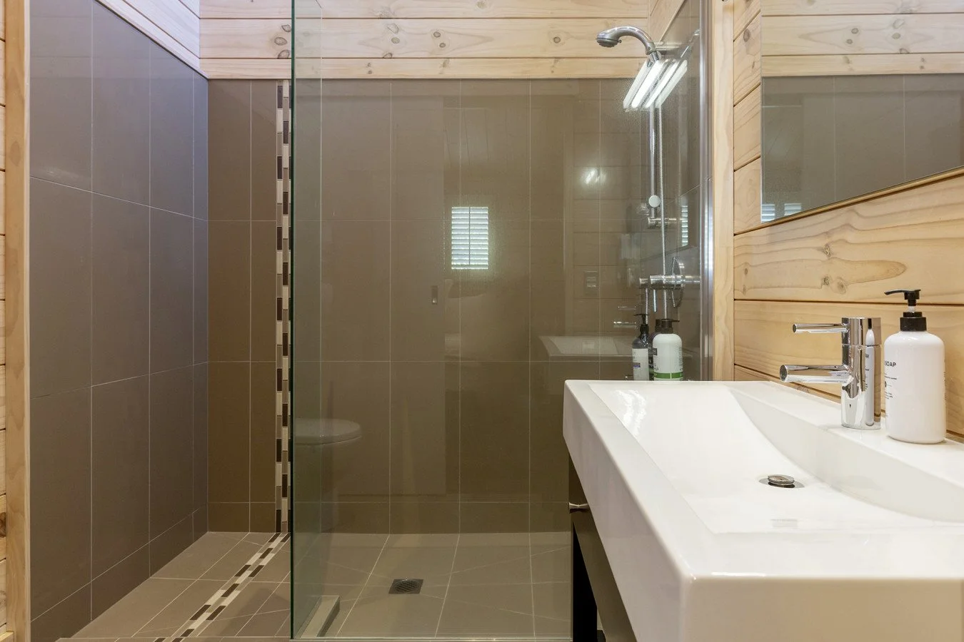 Modern bathroom with a walk-in shower with beige tiles and a glass door, a white sink with a chrome faucet, a mirror, and wooden wall paneling. There are soap and lotion dispensers on the sink and toiletries on a shelf in the shower.